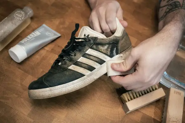 Händer rengör en smutsig Adidas Gazelle-sneaker med en trasa. Bredvid ligger rengöringspasta och borstar, redo att impregnera sneakers.