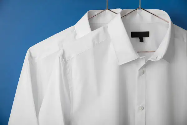 Två vita skjortor hänger på galgar mot en blå bakgrund. De ser ut som om de precis kommit från en **dry cleaner**, perfekt strukna och redo att bäras.